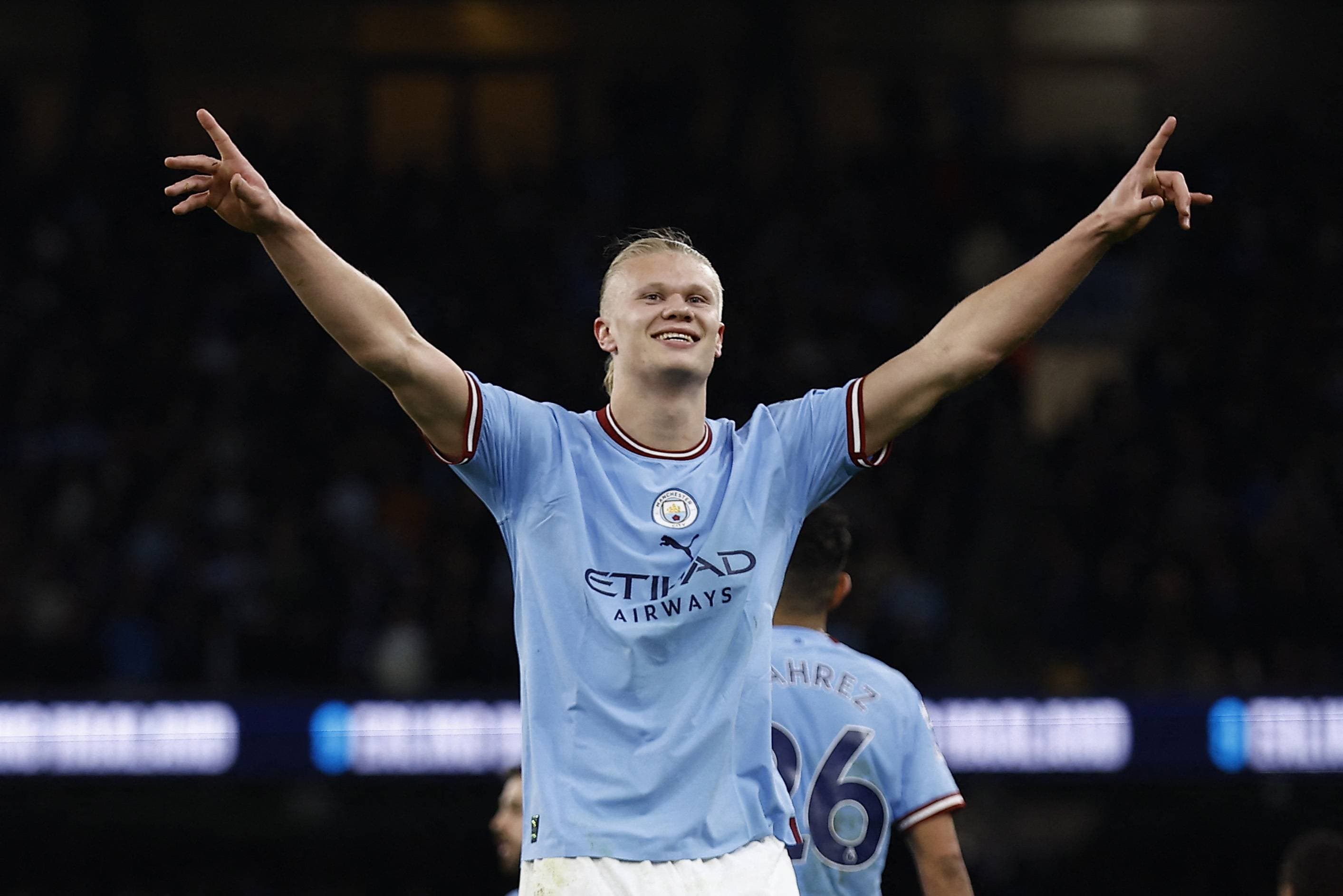 Erling Haaland celebrating with both arms raised at a floodlit Etihad Stadium